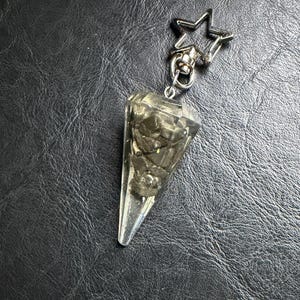 May include: A clear, faceted crystal pendulum with a silver-colored star-shaped clasp. The pendulum is filled with a dark, textured material. The crystal is cone-shaped and tapers to a point. The clasp is attached to a silver-colored ring.