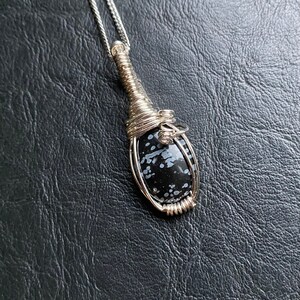 May include: A silver pendant necklace featuring a teardrop-shaped, black and white speckled stone. The stone is encased in a wire-wrapped setting and hangs from a delicate silver chain. The pendant is displayed against a dark background.