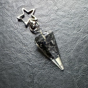 May include: A black crystal pendulum with a silver-colored metal tip and a star-shaped keychain attachment. The crystal is faceted and appears to be filled with dark inclusions. The background is a textured, dark gray surface.