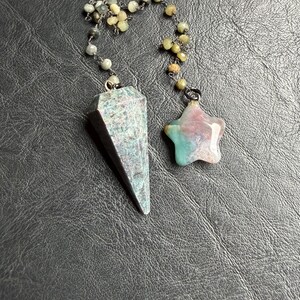 May include: A pendulum and a star-shaped charm, both suspended by beaded chains. The pendulum is a faceted, teal and gray stone. The star charm is pink, teal, and white. The beads are a mix of gray and yellow-green.
