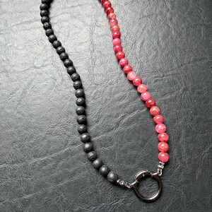 May include: A necklace featuring a combination of black lava beads and faceted red and pink gemstones. The necklace is connected with a silver-colored ring clasp. The beads are strung together, creating a two-toned design.