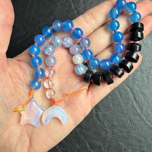 May include: A handmade beaded necklace featuring blue, clear, and black beads. The necklace includes a star and crescent moon charm. The beads are strung on pink and orange string. The necklace is displayed on a hand.