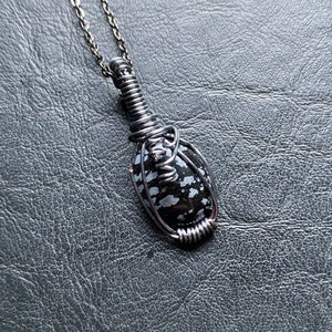 May include: A handcrafted pendant necklace featuring a black and white snowflake obsidian stone wrapped in dark metal wire. The pendant hangs from a delicate chain. The stone has a speckled pattern.