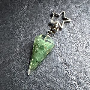 May include: A green crystal pendulum with a star-shaped clasp. The pendulum is cone-shaped and filled with green stones. The metal clasp is silver-colored. The background is a dark, textured surface.