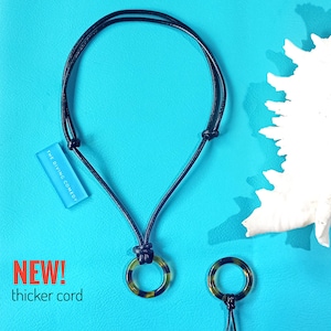 May include: A black cord necklace with a tortoiseshell pendant. The pendant is a ring with a black cord running through it. The necklace is on a blue background with a white coral piece. The text "THE DIVING COMEDY" is on a blue rectangle.