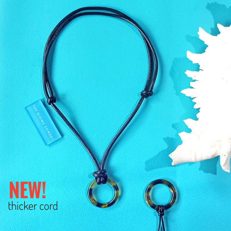 May include: A black cord necklace with a tortoiseshell pendant. The pendant is a ring with a black cord running through it. The necklace is on a blue background with a white coral piece. The text "THE DIVING COMEDY" is on a blue rectangle.