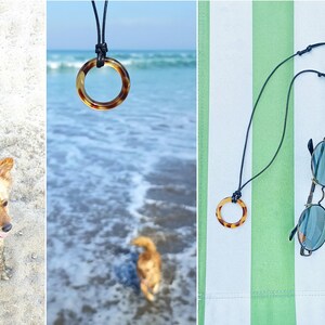 May include: A black leather cord necklace with a brown tortoiseshell ring pendant. The necklace is lying on a green and white striped beach towel with a pair of round sunglasses.