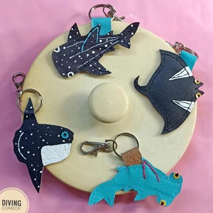 Könnte beinhalten: Handgefertigte Leder-Schlüsselanhänger mit Designs von Meereslebewesen, darunter Walhai, Mantarochen, Hammerhai und Mondfisch. Jeder Schlüsselanhänger hat einen Metallverschluss und eine Lederschlaufe. Hintergrund: hellgelber Kreis auf rosa Oberfläche.