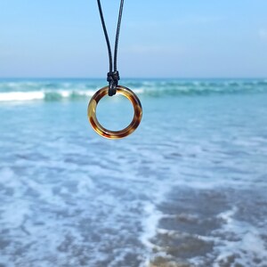 May include: A tortoiseshell ring pendant on a black cord necklace hangs against a backdrop of blue ocean waves.