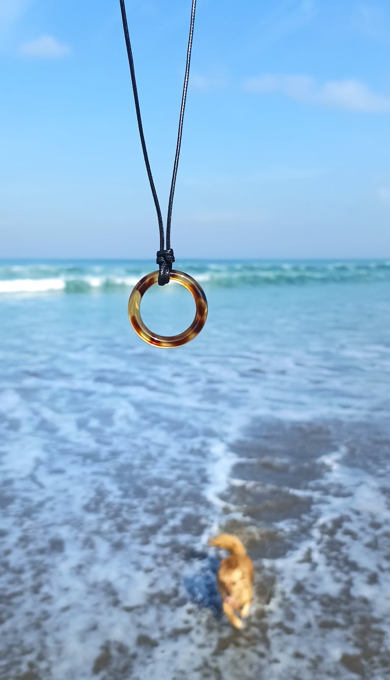 May include: A tortoiseshell ring pendant on a black cord necklace hangs against a backdrop of blue ocean waves.