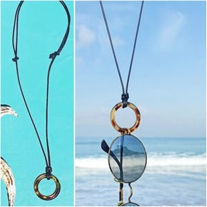 May include: A black cord necklace with a tortoiseshell ring and a pair of round sunglasses hanging from it. The necklace is photographed against a blue background.