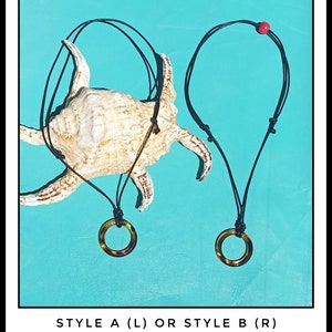 May include: Two necklaces with black cords and round, brown, tortoiseshell pendants. The necklaces are displayed on a seashell against a turquoise background. The text "STYLE A (L) OR STYLE B (R)" is displayed below the necklaces.