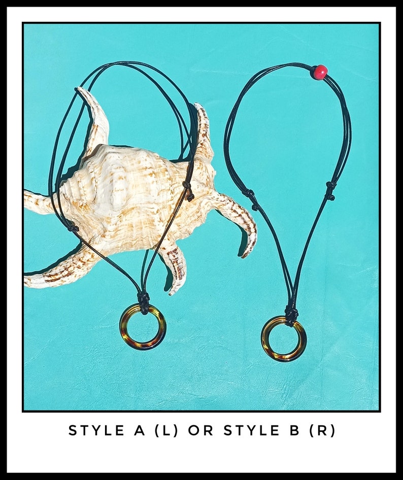May include: Two necklaces with black cords and round, brown, tortoiseshell pendants. The necklaces are displayed on a seashell against a turquoise background. The text "STYLE A (L) OR STYLE B (R)" is displayed below the necklaces.