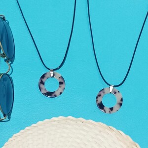 Op de afbeelding: Twee kettingen met cirkelvormige hangers met een schildpadpatroon, hangend aan zwarte koorden. De hangers zijn bevestigd aan zilverkleurige metalen bevindingen. Een zonnebril en een strohoed zijn gedeeltelijk zichtbaar tegen een helderblauwe achtergrond.
