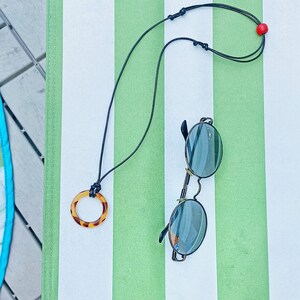 May include: A black cord necklace with a brown tortoiseshell pendant and a red bead. The necklace is lying on a striped green and white surface with a pair of round sunglasses.
