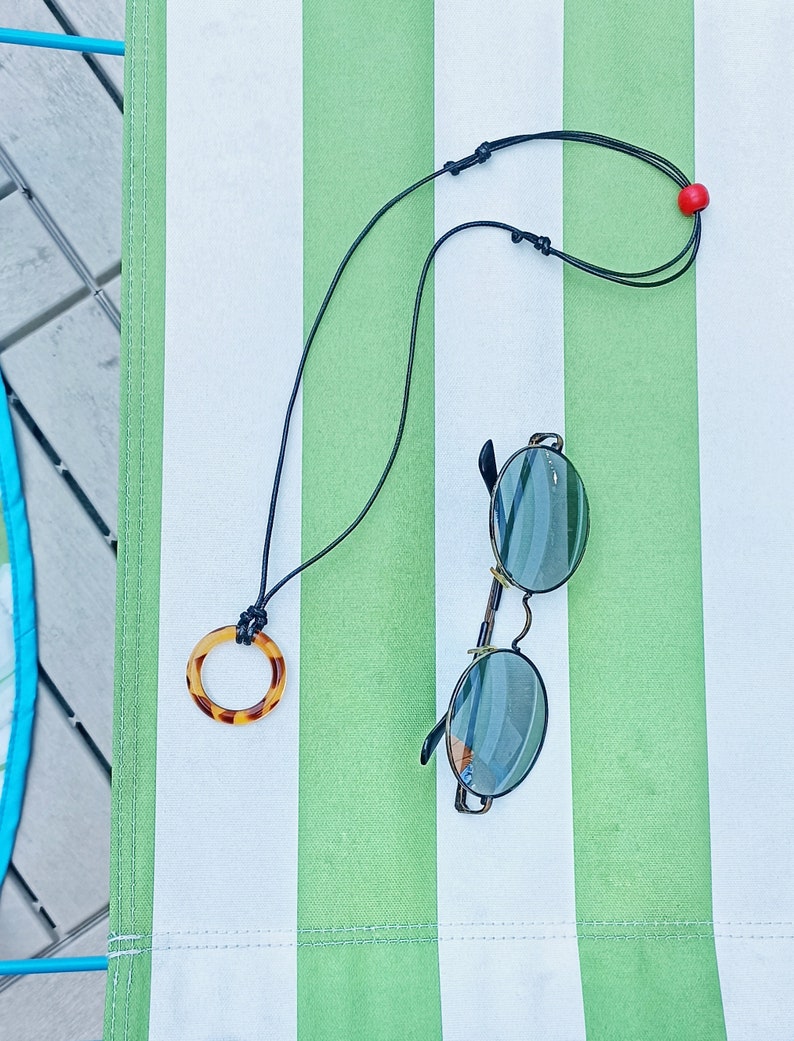 May include: A black cord necklace with a brown tortoiseshell pendant and a red bead. The necklace is lying on a striped green and white surface with a pair of round sunglasses.
