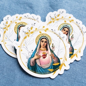 Puede incluir: Tres pegatinas circulares con una ilustración de la Virgen María con velo azul, prenda rosa y un corazón en llamas. Cada pegatina está enmarcada por un borde circular de color dorado con delicados detalles de hojas doradas y un halo.