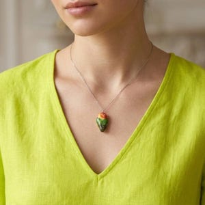Pode incluir: Um colar de prata com um pingente em forma de pássaro colorido. O pingente apresenta tons de verde, laranja e vermelho. A mulher na imagem está usando uma camisa verde limão com decote em V.