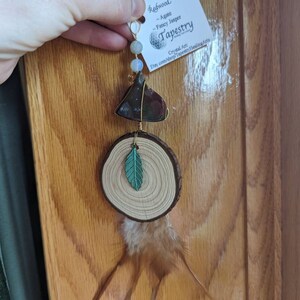 May include: A handmade dreamcatcher with a wooden slice, a green leaf, a brown and red agate, and a brown feather. The dreamcatcher is hanging from a string with white and blue beads. The tag reads "Redwood - Agate - Fancy Jasper Tapestry Crystal Art Etsy.com/shop/TapestryHealingArts"