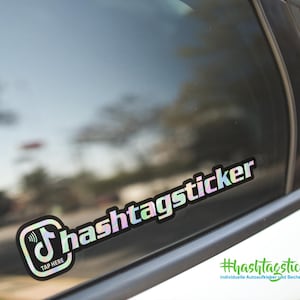 Könnte beinhalten: Ein holografischer Aufkleber auf einer Autoscheibe mit dem Wort "hashtagsticker" in einer stilisierten Schriftart. Der Aufkleber enthält ein Musiknoten-Symbol mit den Worten "TAP HERE". Der Hintergrund zeigt eine verschwommene Straßenszene.