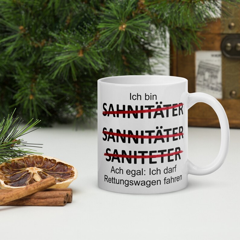 以下が含まれることがあります： 白いセラミック製のマグカップに、黒い文字で「Ich bin Sahnitater Sannitater Saniteter Ach egal: Ich darf Rettungswagen fahren」と書かれています。「Sahnitater」、「Sannitater」、「Saniteter」という単語は赤い線で消されています。