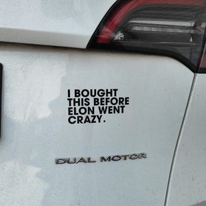 May include: A white Tesla car with a black sticker that reads "I bought this before Elon went crazy." The car also has a black "Dual Motor" emblem.