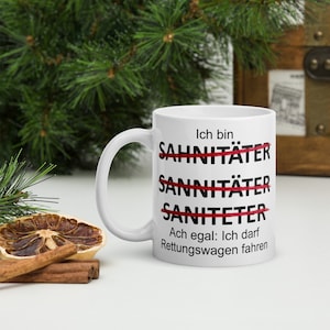 以下が含まれることがあります： 白いセラミック製のマグカップに、黒い文字で「Ich bin Sahnit&auml;ter Sanit&auml;ter Saniteter Ach egal: Ich darf Rettungswagen fahren」と書かれています。「Sahnit&auml;ter」、「Sanit&auml;ter」、「Saniteter」という単語は赤い線で消されています。