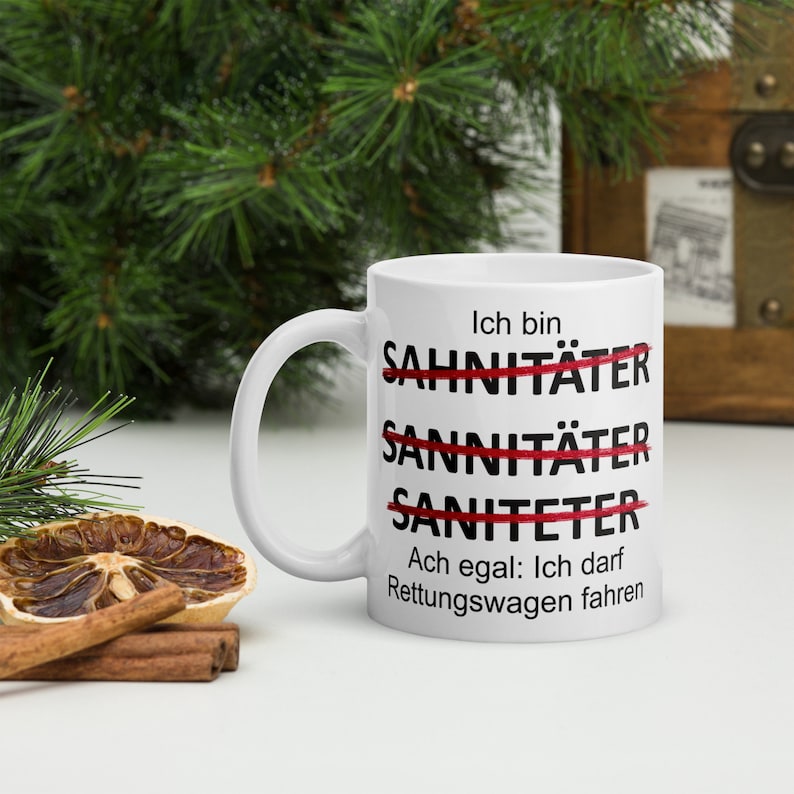 以下が含まれることがあります： 白いセラミック製のマグカップに、黒い文字で「Ich bin Sahnit&auml;ter Sanit&auml;ter Saniteter Ach egal: Ich darf Rettungswagen fahren」と書かれています。「Sahnit&auml;ter」、「Sanit&auml;ter」、「Saniteter」という単語は赤い線で消されています。