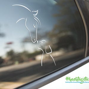 Könnte beinhalten: Weißer Autoaufkleber mit einem Pferd und einem Hund, die sich küssen. Das Design ist eine minimalistische Strichzeichnung des Pferdekopfes und des Kopfes und Körpers des Hundes. Der Aufkleber ist auf der Autoscheibe angebracht.
