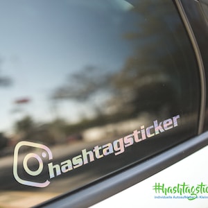 Könnte beinhalten: Eine Autofenster mit einem holografischen Aufkleber, auf dem "hashtag sticker" steht. Der Aufkleber ist ein weißes Kamerasymbol mit einem weißen Kreis darin. Der Text ist in einem weißen, grauen und schwarzen Farbverlauf.