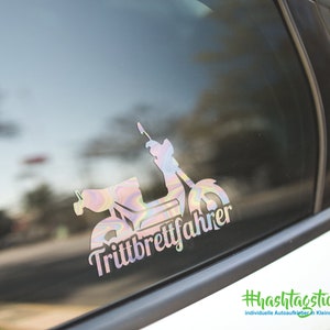May include: A colorful, iridescent sticker of a moped with the text "Trittbrettfahrer" on a car window. The sticker is on a white car with a black trim. The background is blurred.