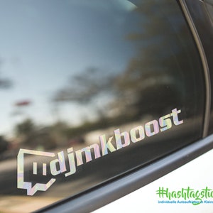 Könnte beinhalten: Ein holografischer Aufkleber mit dem Text "djmkboost" auf einem weißen Autofenster. Der Aufkleber hat die Form eines Twitch-Chat-Icons. Der Text "#hashtagticker.de individuelle Autoaufkleber in Kleinauflage" ist unter dem Aufkleber sichtbar.