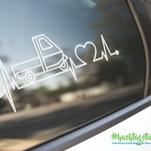 Könnte beinhalten: Weißer Vinyl-Aufkleber mit einem Pickup-Truck, einem Herzen und einer Herzschlaglinie. Der Aufkleber befindet sich auf einer Autoscheibe. Der Text am unteren Rand des Bildes lautet "#hashtagticker.de individuelle Autoaufkleber in Kleinauflage".