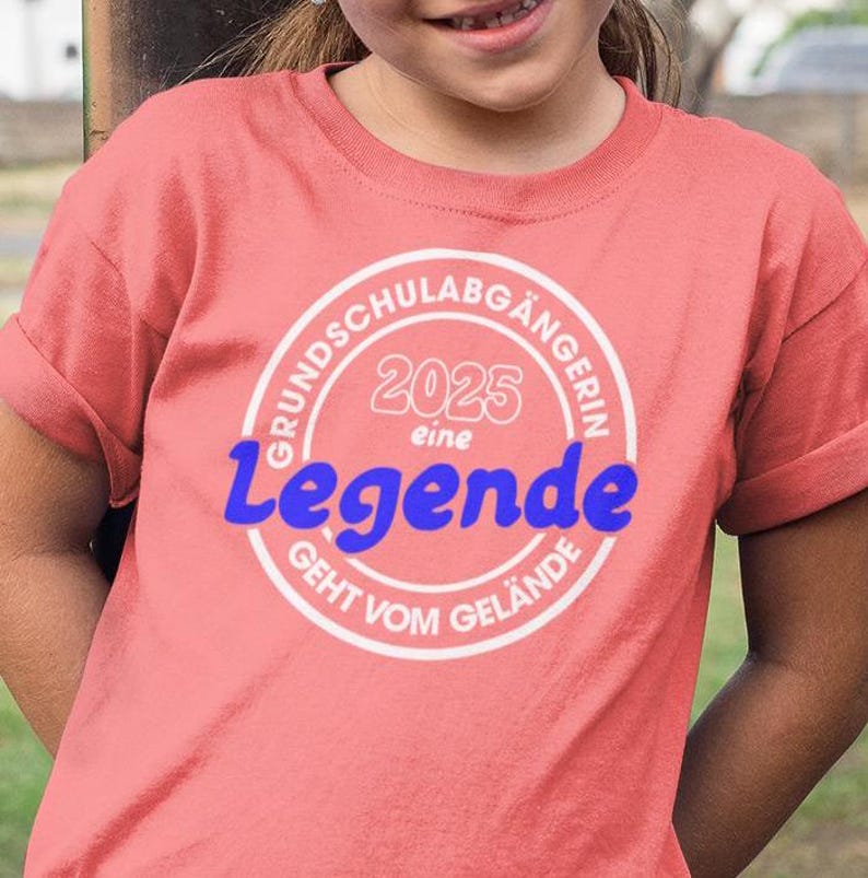 Op de afbeelding: Roze T-shirt met een witte cirkelvormige afbeelding met de tekst "Grundschulabg&auml;ngerin 2025 eine Legende geht vom Gel&auml;nde".