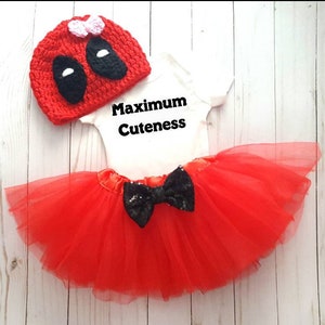 May include: Red and black baby girl outfit with a tutu, a bodysuit that says "Maximum Cuteness", and a crocheted hat with a black mask and pink bow.