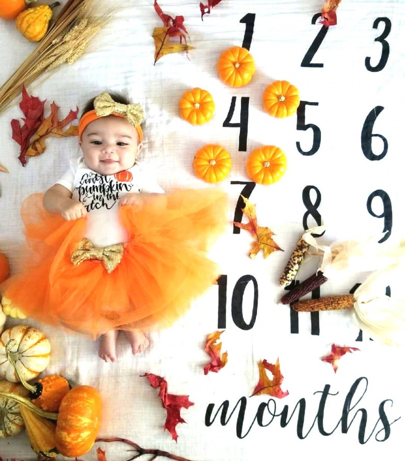 May include: A baby in an orange tutu and headband, with a shirt reading "Cutest Pumpkin in the Patch". The image features decorative pumpkins, autumn leaves, and a milestone blanket marked with numbers and the word "months".