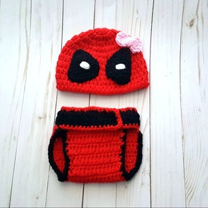 May include: A red and black crocheted baby outfit, including a hat with black eye shapes and a diaper cover. The hat has a small pink flower accent. The outfit is handmade.