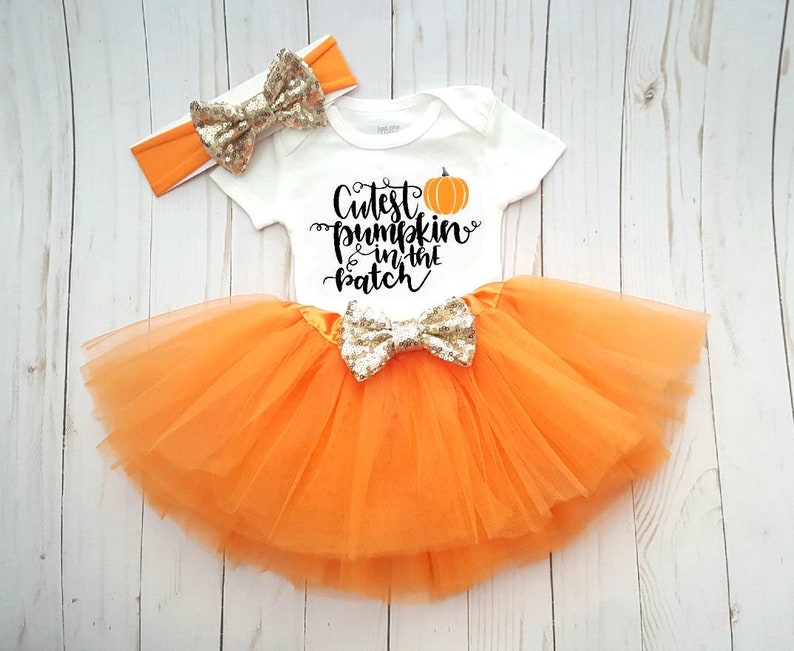 May include: A white baby bodysuit with the text "Cutest pumpkin in the patch" and an orange pumpkin graphic. The outfit includes an orange tutu skirt and a matching headband, both with gold sequin bows. The outfit is designed for a baby.