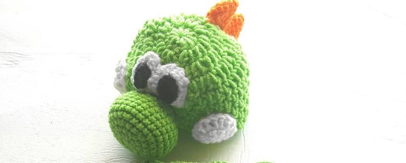 May include: A handmade crochet plush toy shaped like a green dinosaur character. It features a round green head, a prominent green snout, large white eyes with black pupils, and a small orange spike on top. The texture is made of yarn stitches.