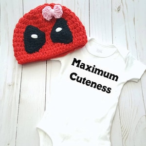 May include: Red and black crocheted hat with a pink bow and white eyes. White onesie with the text "Maximum Cuteness" in black.