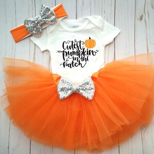 May include: A baby outfit set. It includes a white onesie with the text "Cutest pumpkin in the patch" and a pumpkin graphic. The set features an orange tutu skirt, a silver sequin bow, and an orange headband with a silver sequin bow.