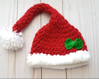 May include: A red crocheted Santa hat with a white fluffy trim and a white pom-pom. A small green bow is attached to the front of the hat. The hat is handmade and suitable for the festive season.
