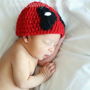 May include: A newborn baby wearing a red and black crocheted hat with a white eye detail. The hat resembles a superhero mask.