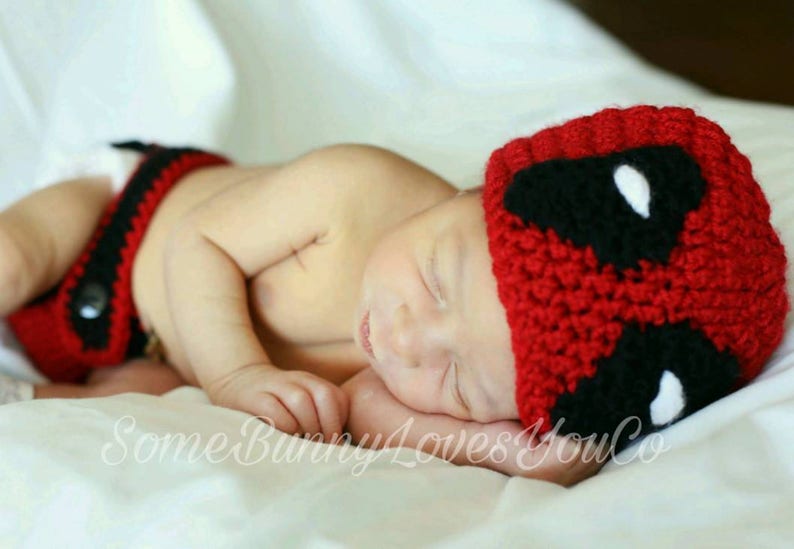 May include: A baby wearing a red and black crocheted hat with a white eye design. The hat is shaped like a superhero mask. The baby is wearing a red and black crocheted diaper cover.