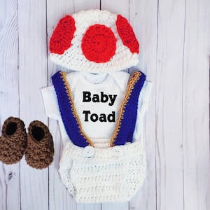 May include: A white crochet hat with red circles, a white bodysuit with the black text Baby Toad, white crochet diaper cover with blue suspenders featuring gold trim, and a pair of brown crochet booties arranged as a complete set.