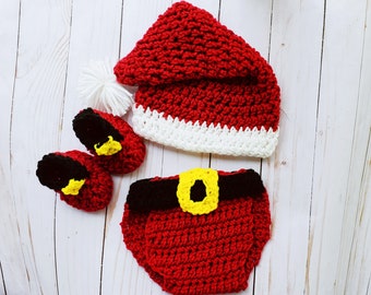 May include: A crocheted baby Santa Claus outfit. It includes a red hat with a white brim and a white pom-pom, red booties with black soles and yellow details, and a red nappy cover with a black belt and yellow buckle.