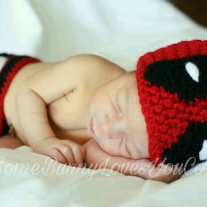 May include: A baby wearing a red and black crocheted hat with a white eye design. The hat is shaped like a character's head. The baby is wearing a red and black crocheted diaper cover.