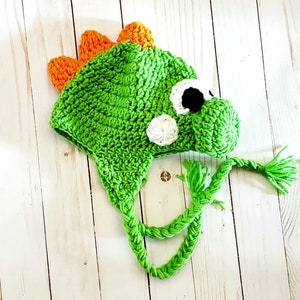 May include: A bright green crocheted hat shaped like a dinosaur, featuring orange spikes along the top, a protruding snout, and white and black circular eyes. The hat includes two long, braided green yarn ties hanging from the sides.