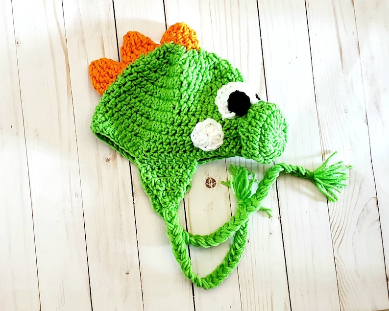 May include: A bright green crocheted hat shaped like a dinosaur, featuring orange spikes along the top, a protruding snout, and white and black circular eyes. The hat includes two long, braided green yarn ties hanging from the sides.