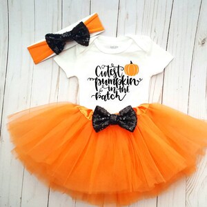 May include: A white baby bodysuit with the text "Cutest pumpkin in the patch" and an orange pumpkin graphic. The outfit includes an orange tutu skirt and a matching headband, both with black sequin bows. The outfit is orange and black.